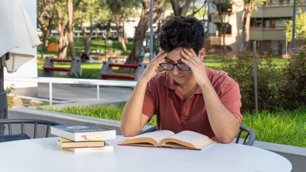 A young student studying at a table in an outdoor university campus setting, concentrating on his books.